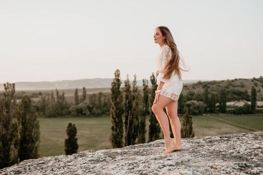 Romantic beautiful bride in white dress posing with sea and mountains in background. Stylish bride standing back on beautiful landscape of sea and mountains on sunset