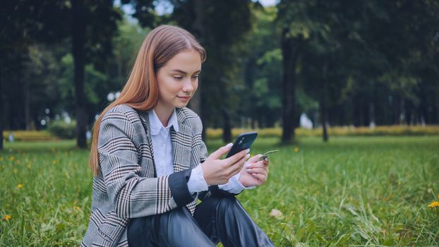 A young girl makes a purchase with her phone and bank card in the park, sitting on the grass