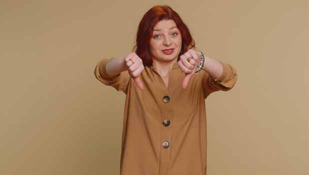 Dislike. Upset unhappy redhead woman in blouse showing thumbs down sign gesture, expressing discontent, disapproval, dissatisfied, dislike. Young adult girl. Indoor studio shot on beige background