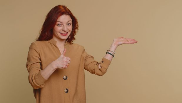 Lovely woman in blouse showing thumbs up and pointing empty place, advertising area for commercial text, copy space for goods promotion. Young redhead girl. Indoor studio shot on beige wall background