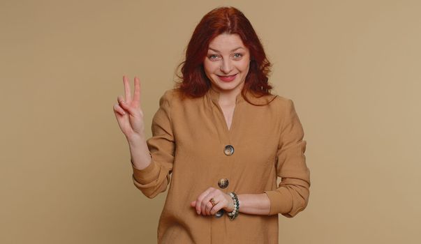 Hipster redhead woman in blouse showing victory v sign, hoping for success and win, doing peace gesture, smiling with kind optimistic expression. Young adult girl isolated on beige studio background