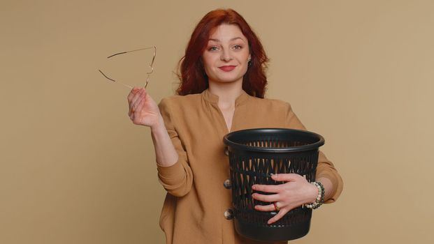 redhead woman taking off, throwing out glasses into bin after medical vision laser treatment therapy surgery, looking smiling at camera. Young ginger girl isolated alone on beige studio background
