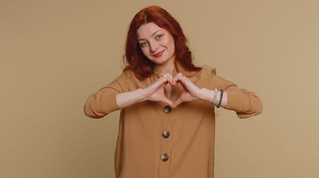 Woman in love. Smiling redhead woman 20s in blouse makes heart gesture demonstrates love sign expresses good feelings and sympathy. Young pretty adult girl isolated alone on beige studio background