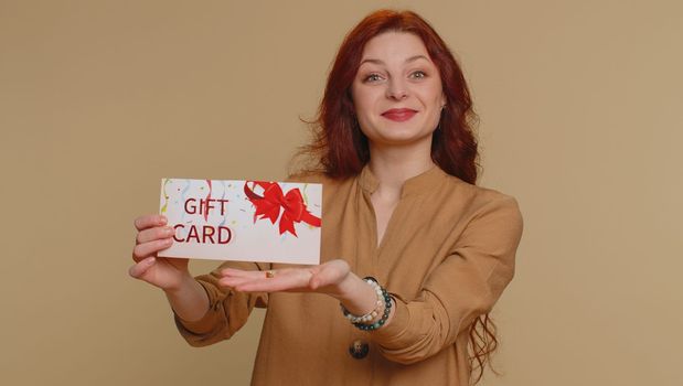 Young woman in blouse showing pointing on gift discount certificate voucher coupon for store holidays sale. Redhead girl with shopping christmas surprise card isolated alone on beige studio background