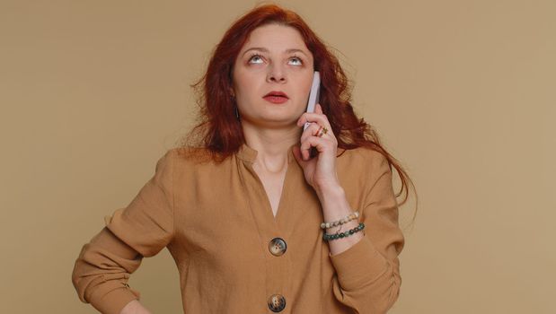 Redhead woman tired sleepy freelancer talking on mobile phone with friend, making online conversation. Disinterested sad young girl having annoyed boring talk on smartphone on beige studio background