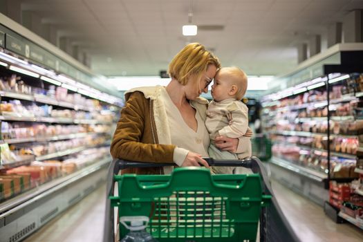 Mother pushing shopping cart with her infant baby boy child down department aisle in supermarket grocery store. Shopping with kids concept