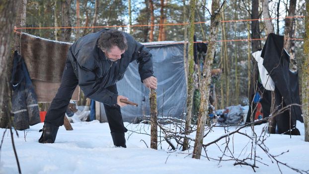 A homeless man is sawing a tree in the woods in front of his cabin