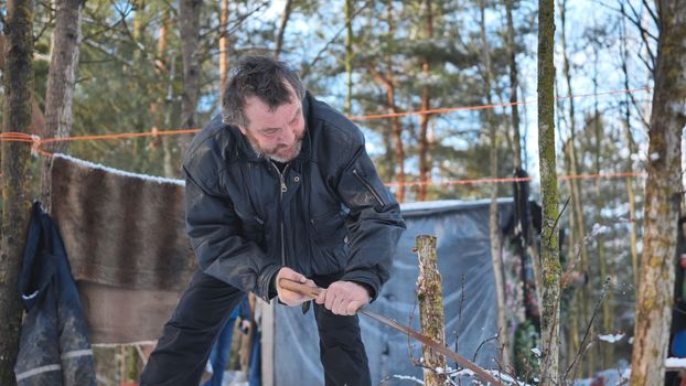 A homeless man is sawing a tree in the woods in front of his cabin