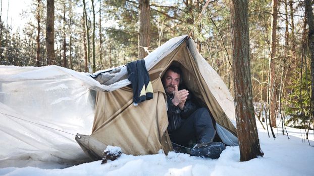 A homeless man climbs in and out of a tent in the woods in winter