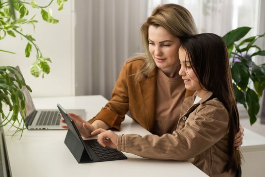 Teen child daughter studying at home in kitchen with mom. Teenage school kid girl distance learning virtual online class with mother or tutor helping doing homework together during remote education.
