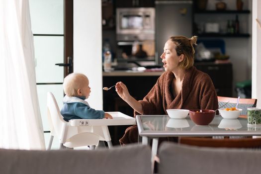 Mother wearing bathrope spoon feeding her infant baby boy child sitting in high chair at the dining table in kitchen at home in the morning.