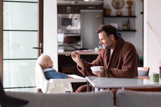 Father wearing bathrope spoon feeding hir infant baby boy child sitting in high chair at the dining table in kitchen at home in the morning.