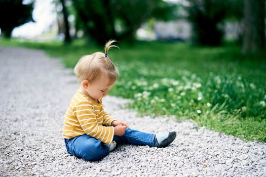 Little girl with a ponytail on her head sits on a gravel path in a green park. High quality photo