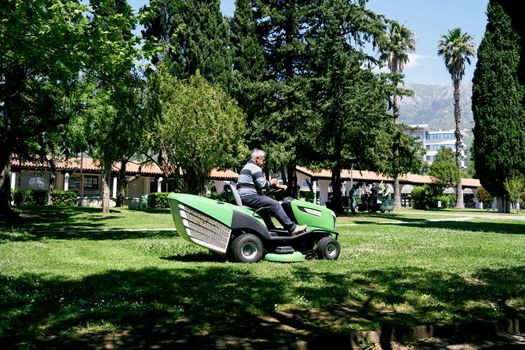 Man rides a green large lawn mower in the park among the trees. High quality photo