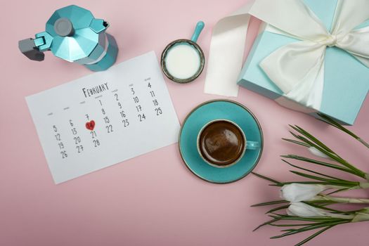 There is a geyser coffee maker on the table, a cup of coffee, a calendar for February and white crocus flowers.