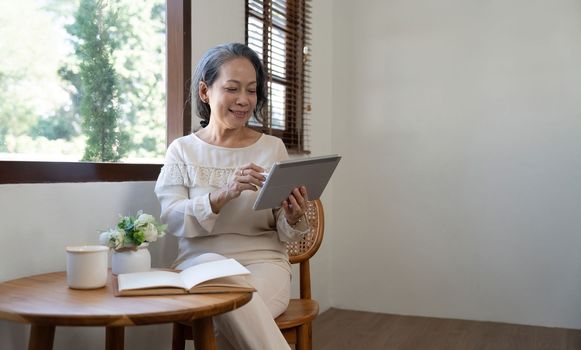 Happy aged Asian old woman sitting by the window and using tablet.