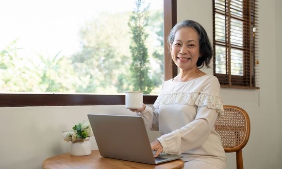 Happy aged Asian old woman working , sipping coffee while using laptop.