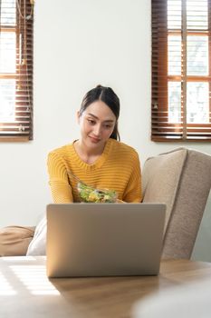 Happy Asian woman eating healthy salad vegetables mix while using her laptop in the living room. work from home.