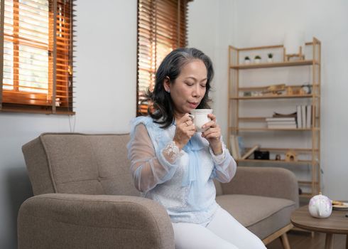 Asian old woman sipping tea in living room at home.