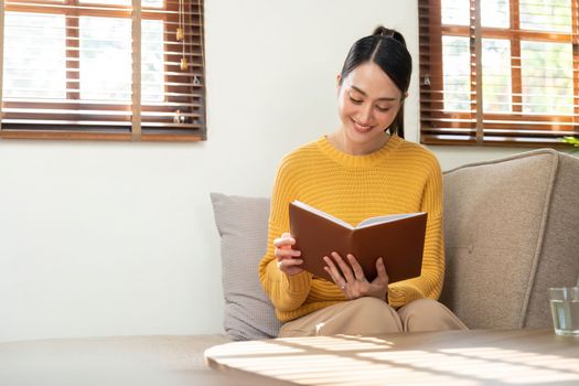 Happy and relaxed Asian woman reading a book on sofa.