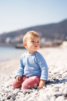 Pensive baby sits, turning to the right, on a pebble beach by the sea against a background of mountains. High quality photo