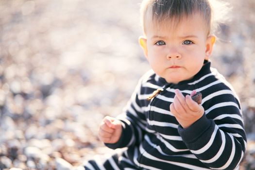 Serious baby in a striped overalls sits on a pebble beach, holding a pebble in his hand. Close-up. High quality photo