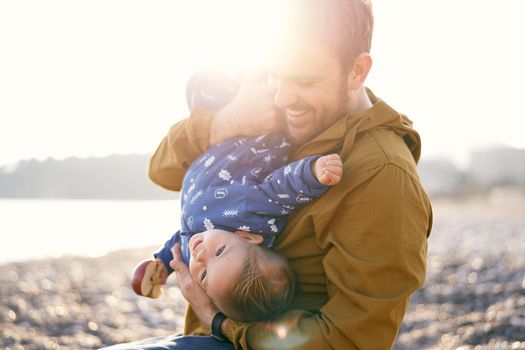 Smiling dad in the bright sunlight hugs a small child in a blue overalls with an apple in his hand to his chest. Close-up. High quality photo