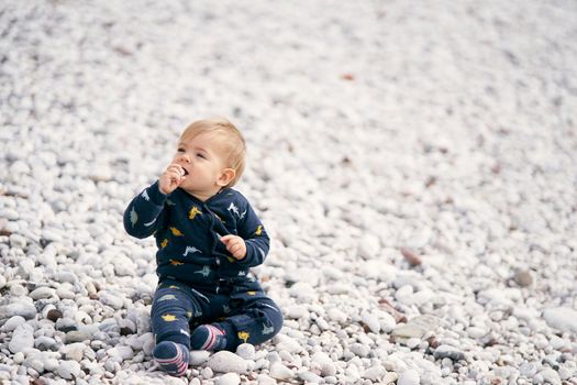 Cute baby in a blue overalls sits on a pebble beach, turning his head to the left and holding a pebble in his hand that pulls into his mouth. High quality photo