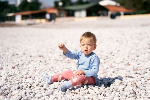 Cute child in a blue blouse and red pants sits on a pebble beach, holding up a pebble in his hand. High quality photo
