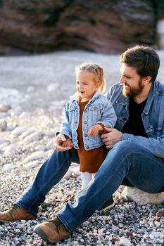 Laughing dad is sitting next to a little laughing girl on a pebble beach. Close-up. High quality photo