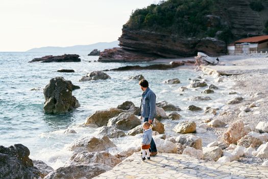 Dad holds the hand of a little girl while standing on a stone path on a pebble beach near the water against a background of green mountains. High quality photo