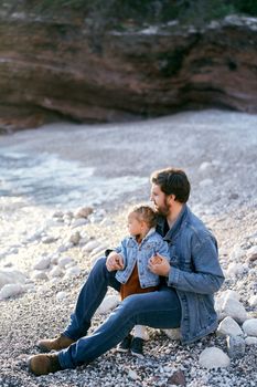 Dad sits on a stone and hugs a little girl on a pebble beach against the backdrop of rocks. Daughter and dad are looking at the sea. Side view. High quality photo