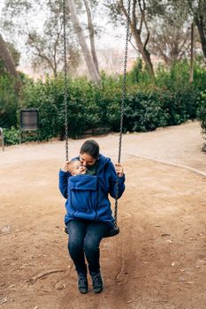 Mom in a blue warm sweater and jeans is swinging on a swing with a tiny baby on her chest. High quality photo