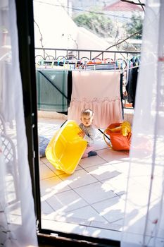Small child sits on the floor on the balcony and holds a yellow bowl in his hands against the background of a clothesline with drying clothes. High quality photo