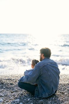 Dad in a denim jacket sits on a pebble beach and holds a little girl in his lap. Back view. High quality photo