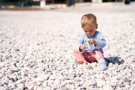 Cute baby in a blue blouse and red pants sits on a pebble beach, bowing his head and holding pebbles in his hands. High quality photo