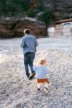 Little girl runs after dad on the pebble beach against the backdrop of rocky mountains. Back view. High quality photo