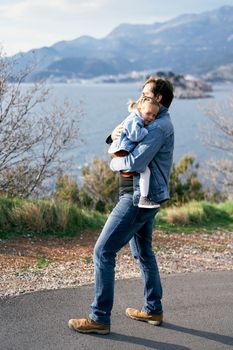 Dad hugs a smiling little girl to his chest, walking along the road against the backdrop of the sea, mountains and the island of Sveti Stefan. High quality photo