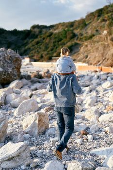 Dad carries a little girl on his shoulders while walking along the pebble beach to green mountains and large boulders. Back view. High quality photo