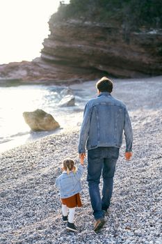 Dad leads a little daughter by the hand, walking along the pebble beach to rocky mountains and the sea. Back view. High quality photo