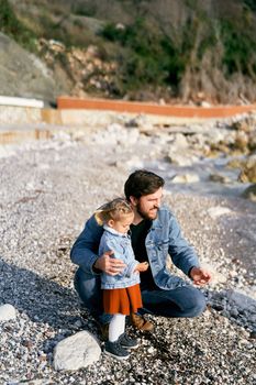 Smiling dad squatted next to the little girl, handing her pebbles from a rocky beach by the water. Close-up. High quality photo