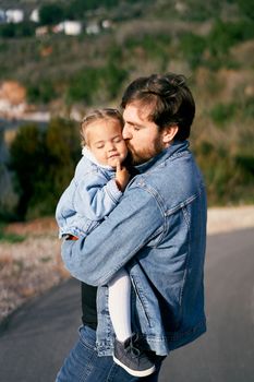 Dad carries and kisses in cheek a little girl in his arms, standing on the road. Close-up. High quality photo