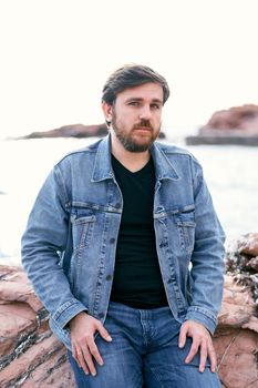 Pensive man in jeans and denim jacket posing sitting on stones against the background of the sea. Close-up. High quality photo