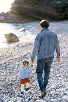 Dad leads a little girl by the hand, walking along the pebble beach against the backdrop of rocky mountains and the sea. Back view. High quality photo