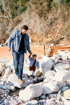 Smiling dad holds the hand of a little girl walking along the stones on a pebble beach against the backdrop of green mountains. High quality photo