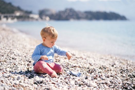 Little kid in a blue blouse and red pants sits on a pebble beach holding a stick in his hand. High quality photo