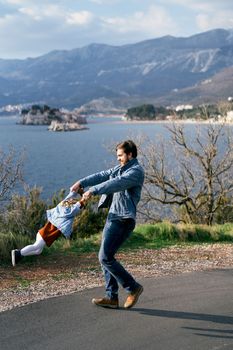 Dad turns his hands around a little girl in a dress and a denim jacket, standing on the road against the background of the sea, mountains and Sveti Stefan island. High quality photo