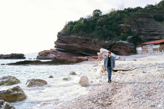Pensive man in jeans and a denim jacket walks along a pebble beach looking at the water against a background of green rocks. High quality photo