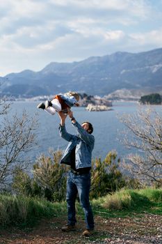 Dad tosses a little girl in a dress and denim jacket, standing on the coast against the backdrop of the sea, mountains and the island of Sveti Stefan. High quality photo