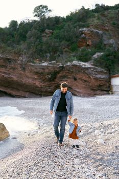 Smiling dad leads a little girl by the hand along a pebble beach against a background of green rocky mountains. High quality photo
